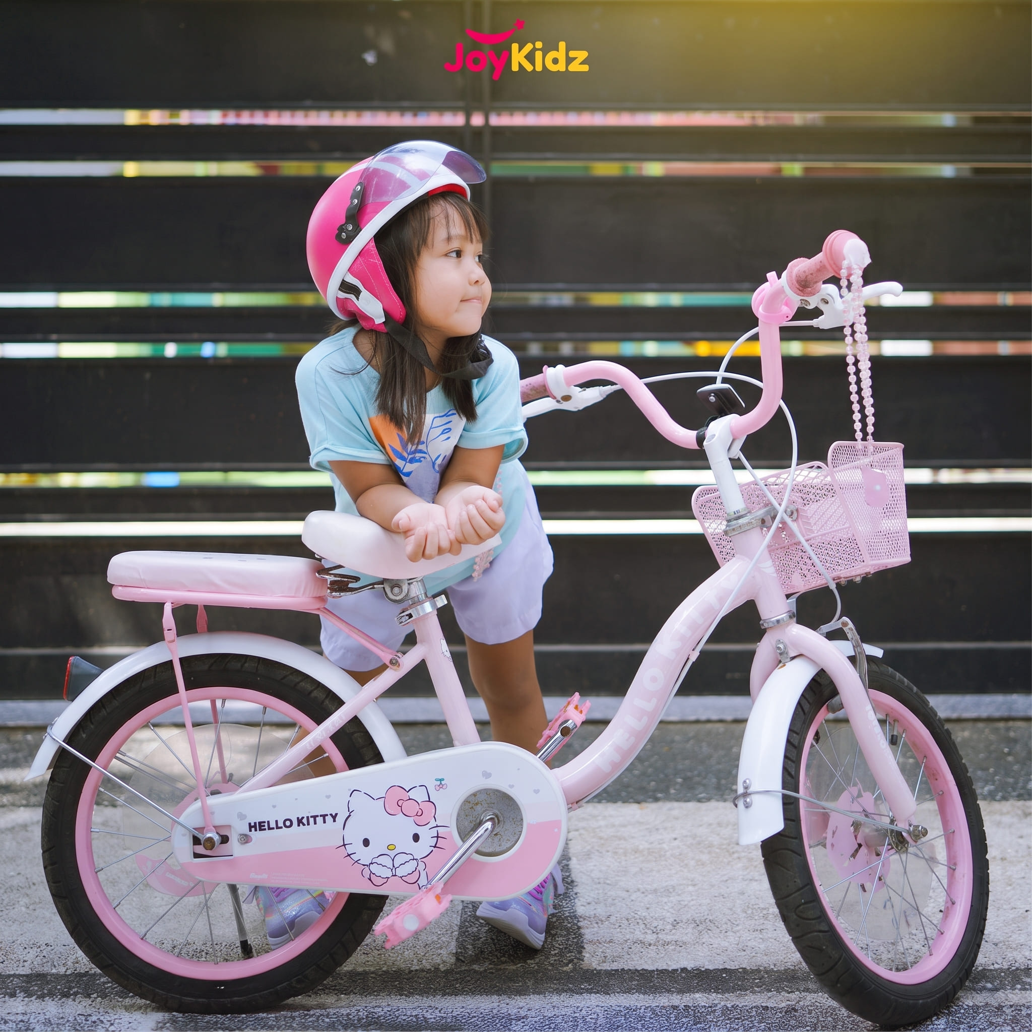 Anak perempuan memakai helm JoyKidz merah muda di samping sepedanya